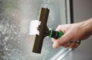 A person uses a green and black squeegee to clean a soapy window.