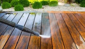 A pressure washer is cleaning a wooden deck. The left side of the deck is wet and clean, while the right side is still dirty. Green shrubs and rocks are in the background.