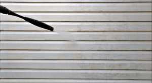 A pressure washer is being used to clean a beige vinyl siding. The left side shows the nozzle spraying water, while the right side shows the siding becoming cleaner, removing dirt and grime.