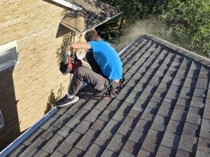 Eavestrough repair Etobicoke: a DT Cleaning technician at the roof edge with a cordless blower, dust blowing off the eavestrough line