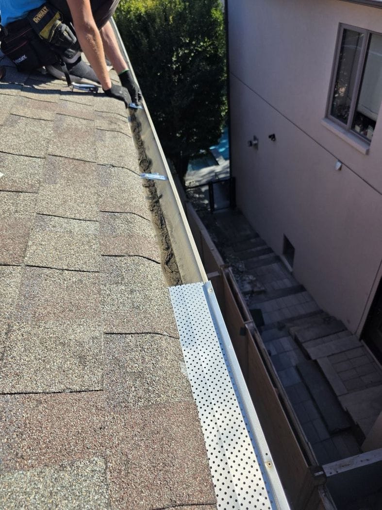 Dark sediment in the trough behind a perforated leaf guard panel, a technician's gloved hands at a bracket, Etobicoke