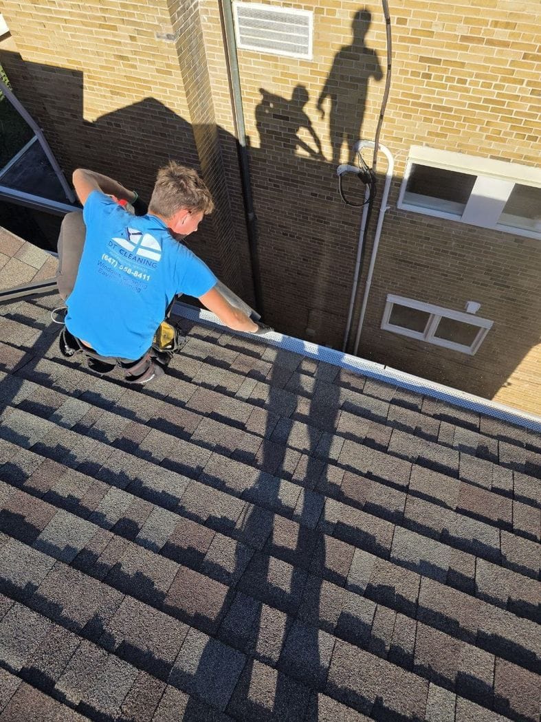A DT Cleaning technician crouched at the roof edge with one hand on the perforated leaf guard of an Etobicoke house