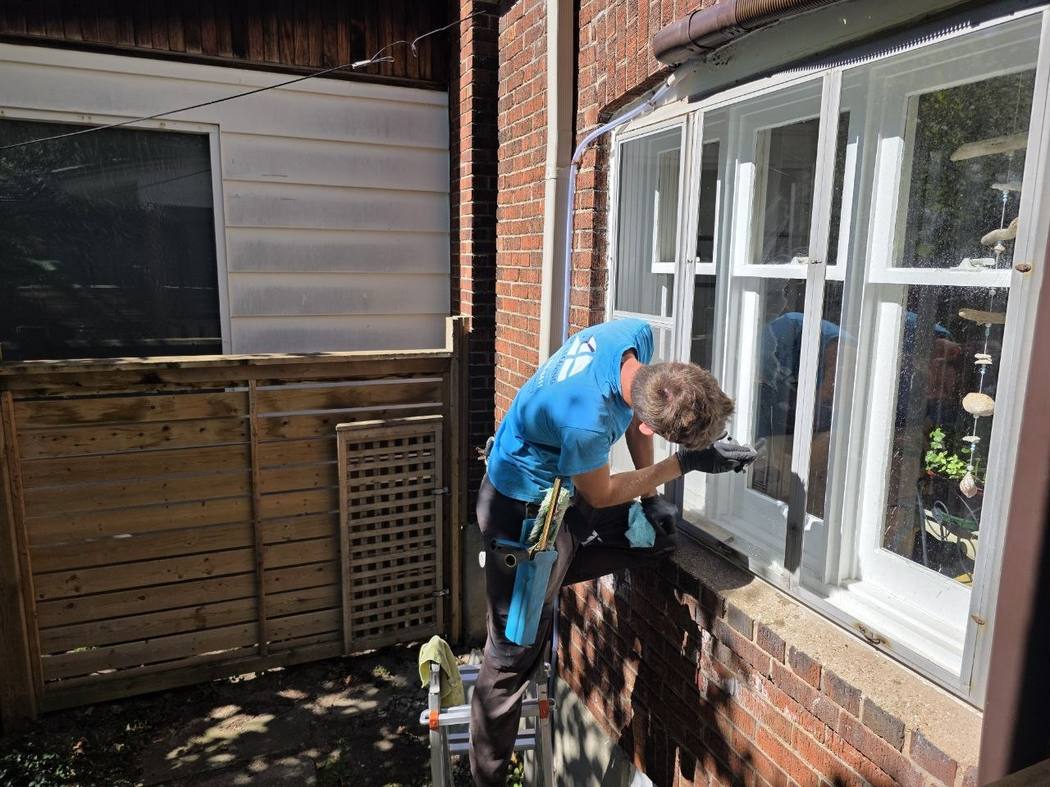 Technician on a ladder at a storm window, Etobicoke A DT Cleaning technician on a ladder with one knee on the sill, working at the lower part of a storm window frame in Etobicoke