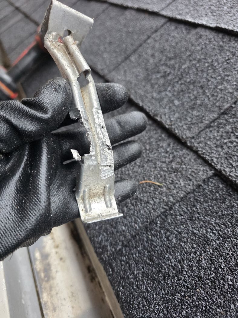 Downspout repair Etobicoke: a gloved hand holding a removed gutter bracket with its strap cut through, over an asphalt shingle roof