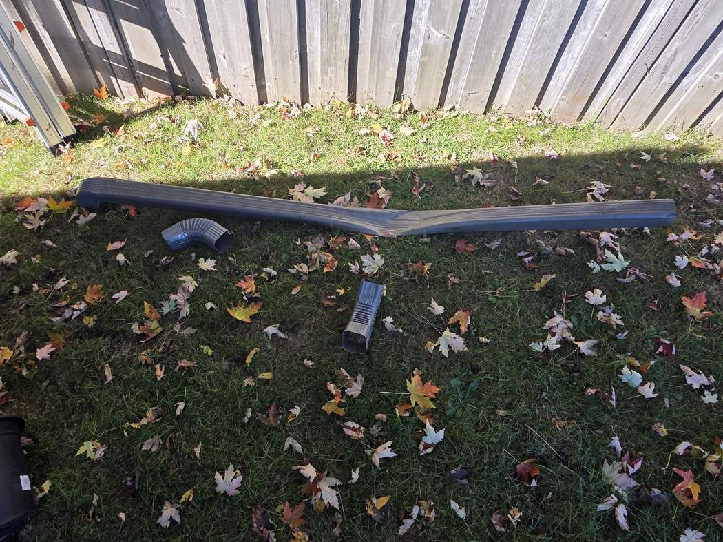 The damaged downspout the crew replaced, Etobicoke The old grey downspout lying bent and kinked on the grass by a wooden fence, with a loose elbow beside it, Etobicoke