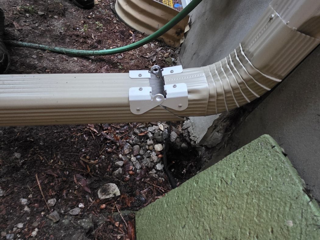Downspout repair Etobicoke: a hinged aluminum downspout extension at ground level beside the foundation of a house, October 2026