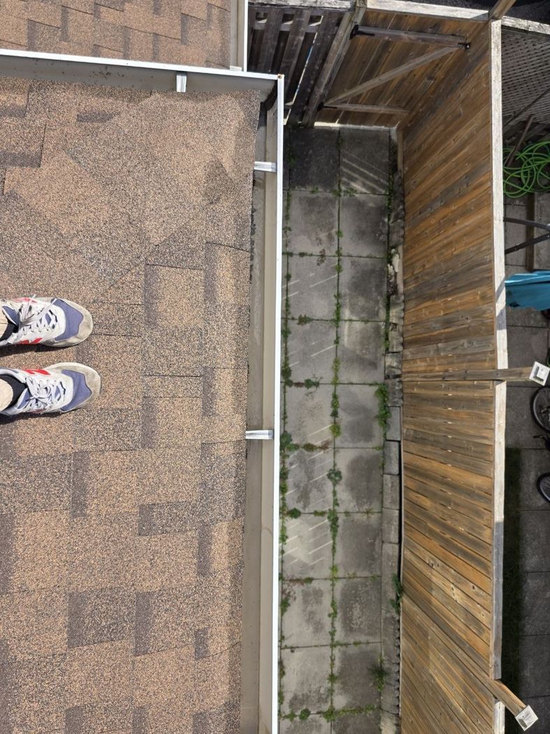 Eavestrough repair Mississauga: a corner of the eavestrough with brackets across it on a shingle roof, a paver walkway and wooden fence below