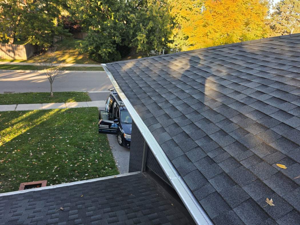 Gutter guards along the roof edge, Etobicoke White perforated gutter guards along the edge of a dark shingle roof, the DT Cleaning van on the driveway below, Etobicoke
