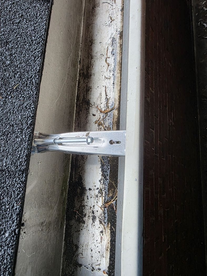 Downspout repair Etobicoke: a new aluminum bracket screwed across the eavestrough, with the shingle roof at left and a brick wall at right