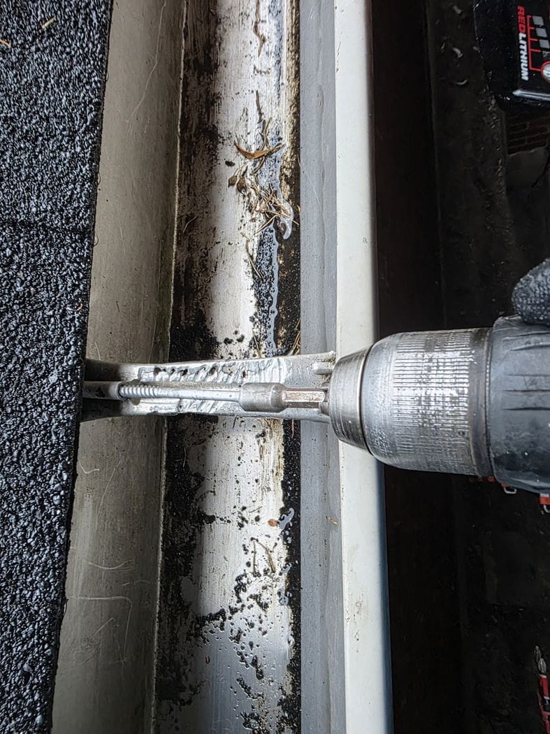 A cordless driver fastening a new bracket across the eavestrough of an Etobicoke house, dark sediment in the trough below