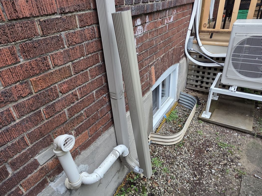 A grey downspout on a brick wall joining a white underground drain pipe with a grated cap at ground level, Etobicoke